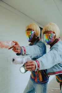Two humans wearing short blond wigs, wearing denim and rainbow masks covering their faces