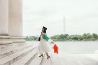 Human walking up the steps of a building with green, white and orange garment over their left shoulder and a graduation cap on