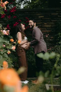 Two humans smiling in colorful floral greenery while one touches the other's baby bump