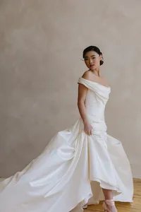 Human dressed in a white wedding gown and standing in front of a mauve backdrop and looking behind them