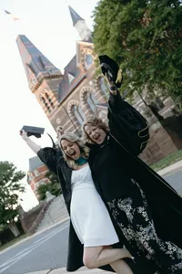 Two individuals holding their graduation caps up in the air and celebrating, one with a white dress and one with a black dress