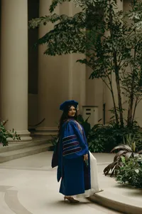 Human in blue graduation grown with black graduation cap looking back at the camera while standing outside with big white pillars and greenery around them