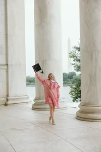 Human in pink holding a graduation cap standing outside with three large marble pillars in the background