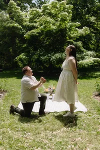 One human proposing to the other, both wearing white, with a white blanket and flowers and a greenery background