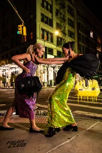 Two humans, one in a sparkly pink and purple dress while the other in a sparkly lime green dress, touching each other's shoulders in city street