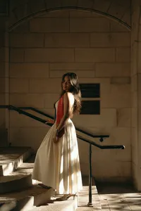 Human walking up the stairs in a building with sunshine hitting their back while they wear a white dress and a red sash