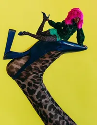 Individual with hot pink hair, neon green shorts, black leopard nylons sitting on a giant stiletto with leopard print stockings and a bright yellow background