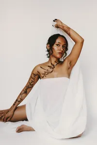 Human wearing white garment with body art from their neck and down to their wrist, with a white background and arm up in the air