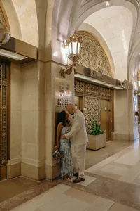 Couple embracing outside elevators with one with a blue dress and flowers and the other in a tan suit