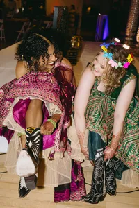 Two individuals smiling and looking at one another while getting their shoes on, both wearing colorful decorated gowns and smiling