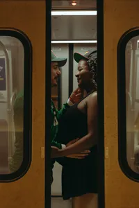 Two humans on the subway, wearing hats, while they both touch the one's baby bump