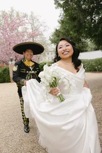Two individuals at their celebration of love, with the background person with a large hat on and the foreground person holding a bouquet of flowers