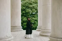 Individual standing in black graduation gown with white dress at the bottom with graduation hat on and greenery in the background