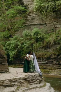 Two humans kissing in front of greenery and cliff area outdoors, while one wears a bridal veil