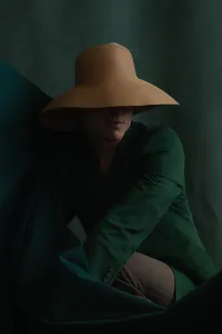 Human with brown hat covering their face with a dark emerald green background and a dark emerald green shirt