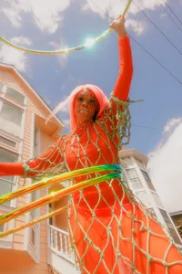 Human with orange wig, orange outfit and sparkle netting-like pattern with blue sky above them