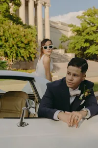 Two humans with one in a bridal gown in the background with sunglasses on, while the other is leaning against the door of the car and looking off to the right side