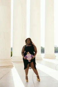 Human standing in a black dress and holding pink graduation cap outside university building