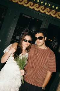 Couple with sunglasses on, one with a striped shirt, the other in a white dress and holding green and white flowers