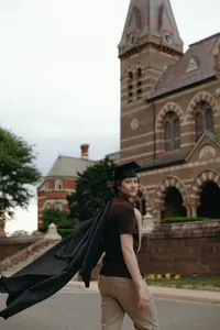 Human with graduation cap on looking over shoulder at camera while walking towards university building