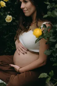 Human with white cropped top and brown pants, cradling baby bump, with yellow flowers surrounding them