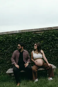 Two humans sitting next to each other with sunglasses on, looking off to the right, one holding their baby bump while they sit against greenery