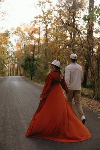 Two humans walking down road in the autumn, holding hands, while one is wearing a red dress and looking back at the camera