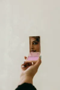 Human holding a makeup mirror with blush and looking into it with a beige background