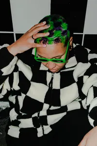 Human in a black and white checkered sweatshirt with neon green sunglasses and neon green hair with their hand on their head