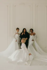Four humans, one in a black dress and three in long, beautiful white dresses with a cream colored background