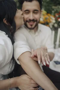 A engaged couple showing off their ring as the soon to be bride kisses her fiancé on the cheek.