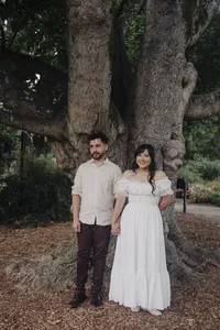 An engaged couple stand behind a large tree holding hands.