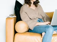Woman sitting comfortably on a tan leather couch working on a laptop, representing a calm and intentional workspace for creative small business owners.