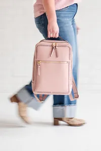 Close-up of a person in cuffed jeans and tan shoes walking while carrying a blush pink backpack with gold zippers.