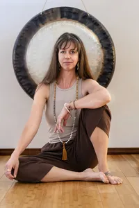 yoga teacher wearing a sleeveless taupe top, brown pants, and a long beaded necklace, seated on a wooden floor in front of a large gong.