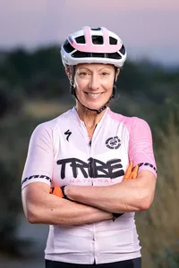 Smiling cyclist wearing a helmet and pink-and-white jersey, standing outdoors with her arms crossed.