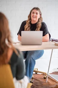 Smiling female nurse practitioner in a black top seated behind an open laptop, talking with another woman across a desk.