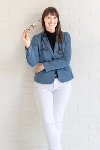 Woman in a blue blazer and white pants with a stethoscope around her neck, holding eyeglasses against a white brick wall.