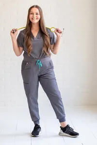 Woman in gray scrubs holding a yellow stethoscope across her shoulders against a white brick wall.