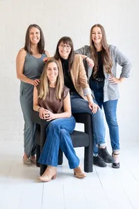 Four smiling women nurse practitioners posing together around a dark armchair against a white brick wall.