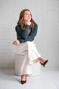 Woman in a dark green top and white pants seated with legs crossed, resting her chin on her hand against a white brick wall.