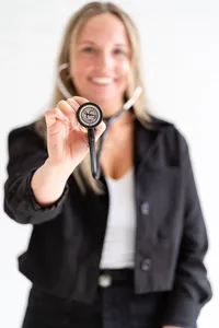 Smiling woman nurse practitioner in a black jacket holding a stethoscope chest piece toward the camera, with her face softly blurred behind it.