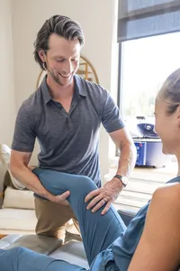 doctor in a gray polo supporting a seated woman’s bent leg with one hand beneath her knee and the other on her shin.