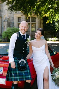 Bride and groom smiling beside a red vintage car at Hatley Castle