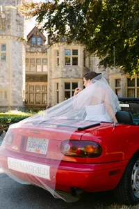 Bride in her wedding dress seated on a red vintage car at Hatley Castle