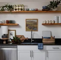 Open shelves styling with colorful dishes, landscape art, and a small lamp aside a kitchen sink.
