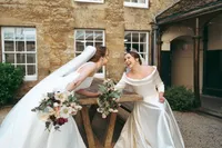Two brides laughing together over a wooden table of bouquets outside the college
