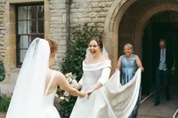 Two brides holding hands and laughing together in a stone archway courtyard, with a bridesmaid holding the dress train