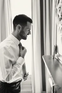 Black and white photo of a groom in profile adjusting his tie beside a window
