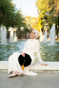 Graduate in a cream hijab and gown seated beside fountains with a graduation cap on her lap